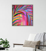 Framed square canvas with vibrant abstract palm leaves in pink, red, green, and blue hues, hanging above a white cushioned chair with a beige pillow and gray throw.