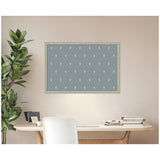 Framed pinboard with a muted blue fabric featuring small, evenly spaced white botanical block print patterns, hanging above a minimalist wooden desk with white chair and green leafy plant.