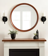 Round wall mirror with a warm wood frame reflecting two window panes, centered above a white mantel decorated with black planters and greenery.
