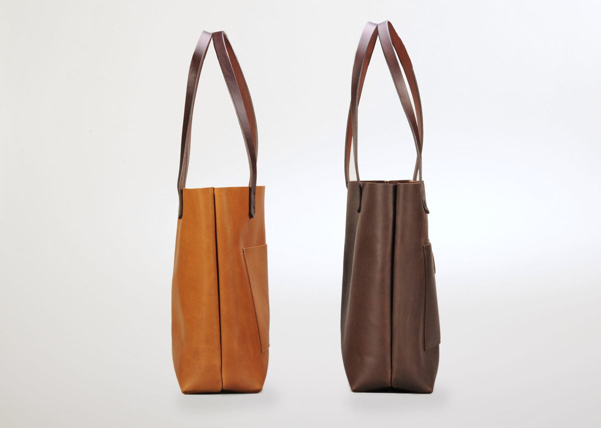 Two leather tote bags shown side by side from their narrow side, one in light brown and the other in dark brown, each with long matching shoulder straps and a front pocket.