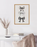 Three detailed black bow illustrations arranged vertically in a light wood frame on a white wall above a white desk with books, vase, and textured blanket.