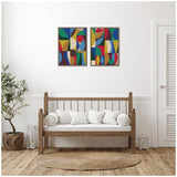 Two framed abstract canvas art pieces featuring bold geometric shapes in blue, red, green, and yellow, hung side by side above a wooden bench with white cushions in a minimalist room.
