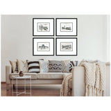 Four vintage Europe framed black-and-white prints arranged in a square above a beige sofa with patterned black, white, and beige pillows and a chunky knit throw blanket.