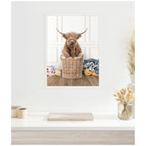 Framed canvas print of a brown highland cow with long curved horns sitting inside a woven laundry basket on wooden floor, surrounded by colorful clothes against white paneled wall.