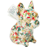 Small ceramic dog-shaped money box decorated with colorful butterfly and red floral patterns, with a 100 currency note inserted in the slot on its back.