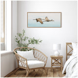 Horizontal framed canvas print of two cranes flying against a soft blue sky, hanging on a white wall above a wicker chair and small wooden side table with a white lamp.