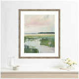 Framed pastel landscape painting of a soft pink sky above green fields with reflective water, hanging on a white wall above a light wood surface with flowers and candle.