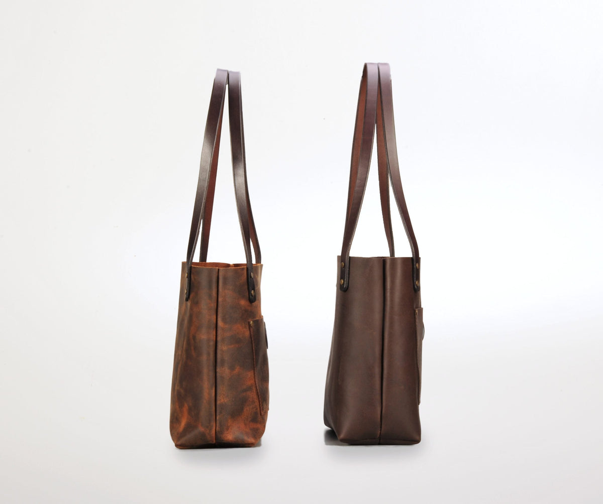 Side view of two brown leather tote bags with long shoulder straps, one in distressed darker brown and the other in smooth dark brown, standing upright against a white background.