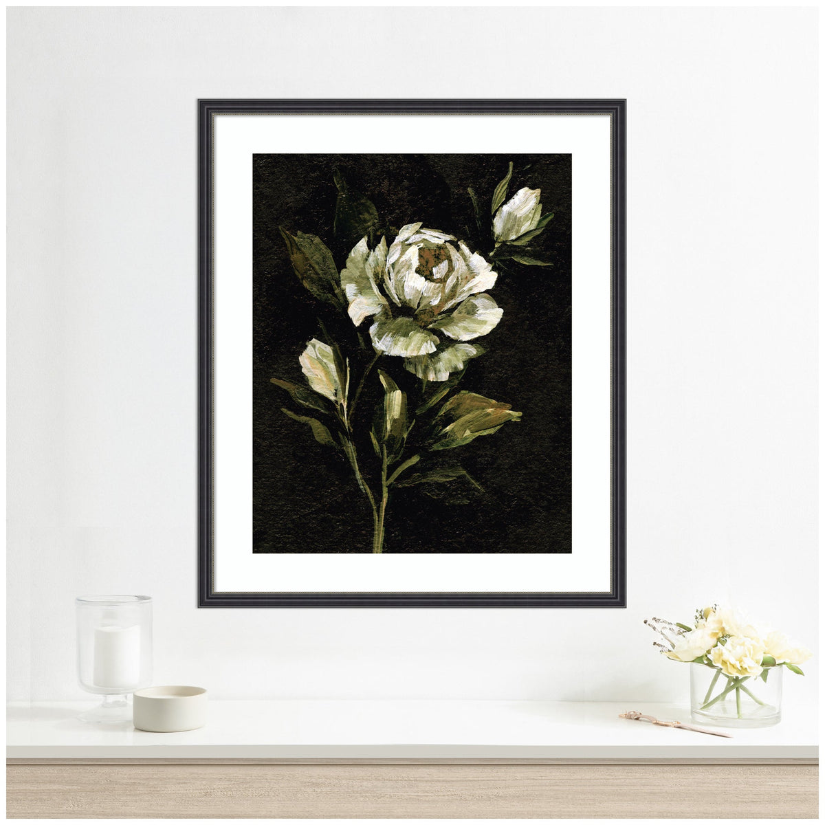 Framed floral artwork featuring a white peony with green leaves on a dark moody background, hanging above a light wooden shelf with glass candle and flower vase.