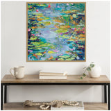 Framed impressionist painting of a colorful water lily pond with vibrant greens, blues, pinks, and yellows, displayed above a wooden table with a mug, books, and a small plant.