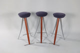 Three modernist bar stools with round purple upholstered seats, angled wooden front legs, and sleek metal rear legs with footrests against a plain light background.