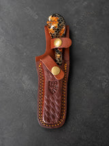 Hand-tooled dark brown leather sheath with yellow stitching, holding a knife with a glossy honeycomb resin handle in amber tones and a brass snap closure.