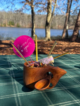 Carved teak kuksa mug with handle and attached wooden spoon, holding colorful items including a pink card with 'love you' script, set on green plaid outdoors near a lake.