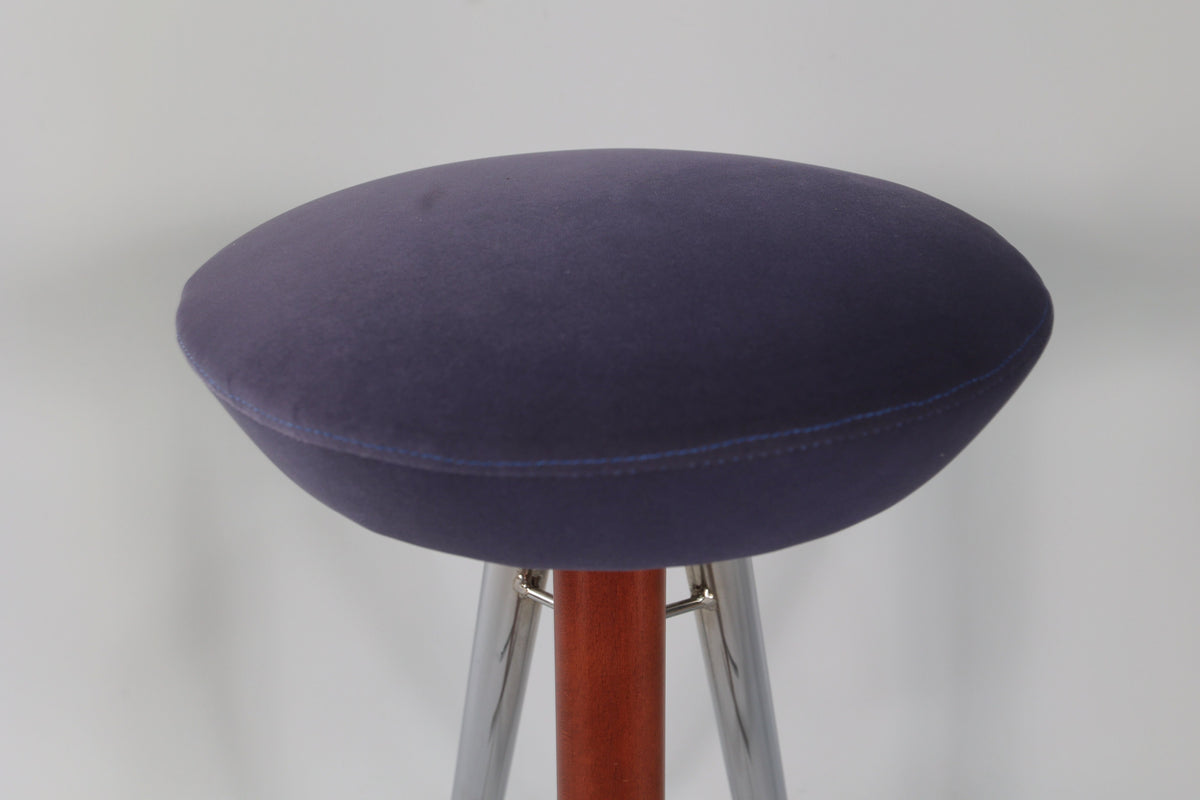 Close-up of a round bar stool seat upholstered in deep purple velvet with neat stitching, supported by a wooden center column and shiny chrome legs.