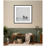 Framed black and white print of a humpback whale breaching the ocean surface with birds flying around, hanging on a beige and sage green wall above a wooden bench.