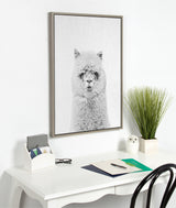Minimalist white desk beneath a framed monochrome alpaca portrait, flanked by a small green plant and an owl-shaped ceramic on a white wall.
