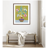 Colorful framed artwork of snowdrop flowers with blue and pink petals in a striped vase on a green and light blue background, hung above a wooden bench with a blanket and open book.