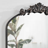 Arched wall mirror with dark ornate scrollwork crest at top, reflecting a room with sheer curtains and potted plants in soft daylight.