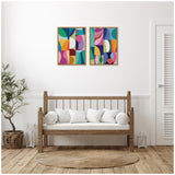 Two framed abstract canvases with colorful geometric shapes in teal, pink, orange, and yellow hang above a wooden bench with white cushions in a minimal room.