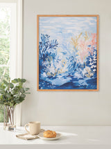 Framed print of an underwater ocean scene with blue, white, and some pink coral shapes against a light blue water background, hanging above a white table with a plant, cup, and croissant.