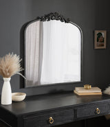 Traditional arch mirror with ornate carved top mounted on a dark gray wall above a black wood dresser with gold ring pulls, styled with a white vase and stacked books.