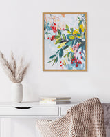 Colorful botanical print with overlapping leaves and flowers in jewel tones of green, yellow, red, and pink, framed in light wood and hung above a white desk with books and a textured throw.