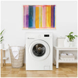 Front-loading white washing machine centered between a woven laundry basket and a white table with laundry items, beneath a colorful abstract canvas with vertical lines.