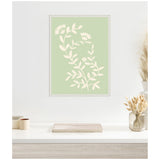 Framed pale green canvas with cream botanical floral pattern hanging on white wall above white table holding closed book, two small cups, and glass vase with dried grasses.