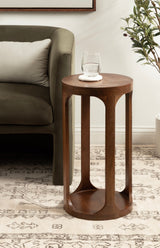 Tall round wooden side table with four open vertical supports and a lower shelf, shown beside an olive green cushioned chair and patterned rug.