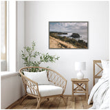 Framed coastal landscape canvas showing rocky cliffs and grassy shore under cloudy sky, hanging above a rattan chair and wooden side table in a bright room.