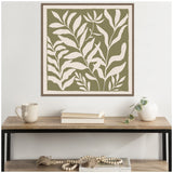 Framed botanical artwork with white abstract leaf shapes on an olive green background, hanging above a wooden console table with neutral decor including a mug, books, and a white vase with trailing greenery.