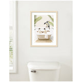 Framed print of a whimsical scene with three penguins bathing in a clawfoot tub surrounded by green palm leaves, mounted on a white wall above a toilet.