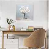 Painting of white daisies in a transparent jar with a soft blue and gray background, hanging centered on a white wall above a wooden desk and beige upholstered chair.