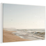 Softly lit sandy beach with gentle waves under a pale blue sky scattered with distant flying birds and faint mountain silhouettes on the horizon.