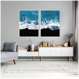 Two square canvas prints side by side showing aerial views of ocean waves crashing onto a black beach with rich blue water and white foam details in a minimalist living room.