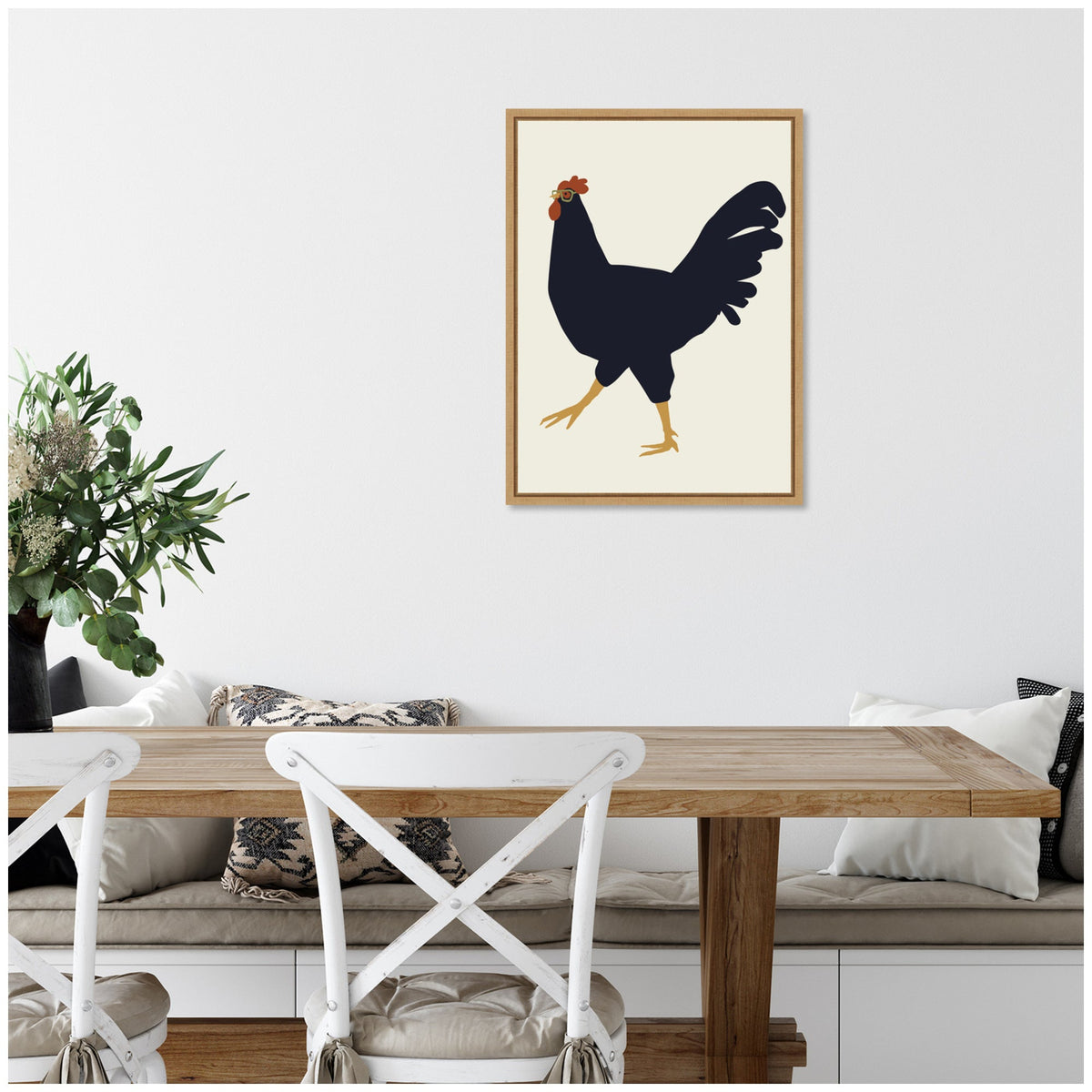 Black silhouette of a rooster with orange comb and yellow legs on a cream background, framed in light wood and hung above a wooden dining table with white chairs.