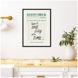 Framed canvas print styled as a guest check with teal and black text that reads 'Take it one day at a time' hanging on a white wall above a light wood countertop with a white pitcher holding green leafy branches.