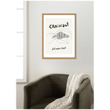 Framed minimalist black and white illustration of a hand holding a croissant with French text saying 'Croissant s'il vous plaît' on a white wall above a dark gray armchair with a knitted blanket.