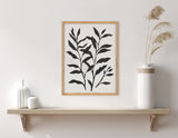 Black botanical silhouettes with elongated leaves on white background, framed in light wood, hanging above a minimalist wooden shelf with white ceramic vases and dried pampas grass.