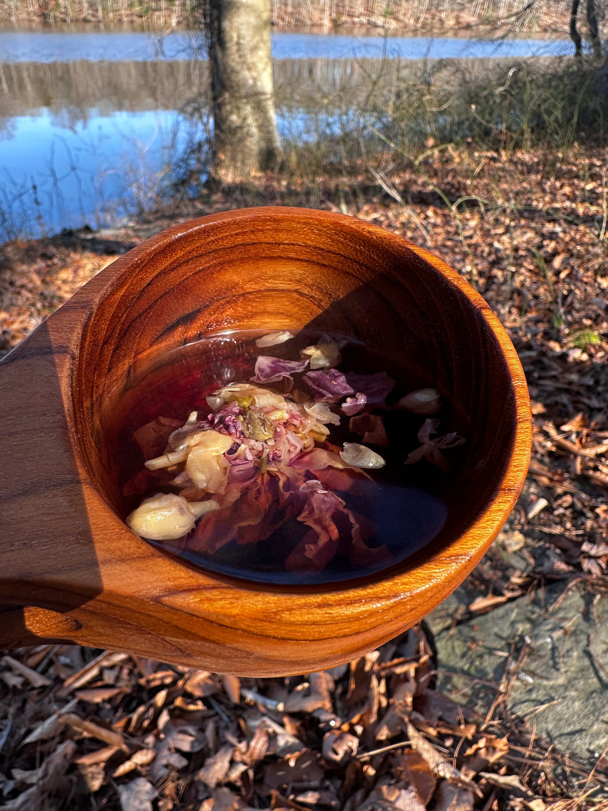 Close-up of a teak kuksa mug with a hanging hole, filled with tea containing floating flower petals, held outdoors near a leaf-strewn ground and a lake in the background.