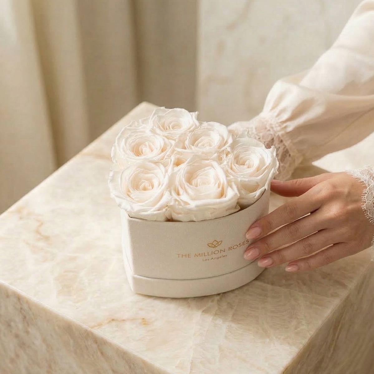 Heart-shaped beige box filled with seven white roses, resting on a cream marble surface with a hand gently touching the box's side.