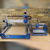 Two blue-framed CNC routers on a wooden surface against a brick wall, one larger with a wide work bed and the other compact with acrylic side panels
