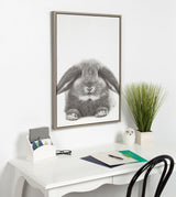 Black and white framed canvas of a fluffy rabbit with long ears, hanging above a white desk with plants and office supplies in a minimalistic room.