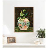 Framed canvas art of a round fishbowl with two orange goldfish, green lily pads, and leaves against a dark green background, displayed on a white wall above a shelf.