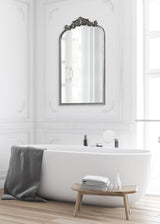 Tall arched mirror with ornate dark decorative top frame reflecting window, mounted on white paneled wall above white freestanding bathtub with gray towel and small wooden bench.