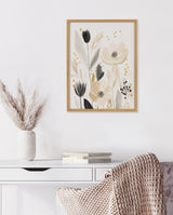 Framed floral print with beige, black, and gray flowers and leaves on a white wall above a white desk with books and a textured beige throw on a chair.