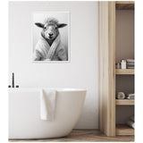 Black and white framed canvas of a sheep wearing a bathrobe, hanging above a white freestanding bathtub with a towel draped over the side in a minimalist bathroom with wood shelving.