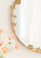 Close-up of an oval mirror with a gold metal frame adorned with detailed floral and butterfly decorations, reflecting soft white curtains beside peach-colored flowers.
