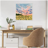 Square framed canvas depicting a sunflower field under a vibrant sunset sky with pink, orange, and blue clouds, hanging above a wooden desk with plants and books.