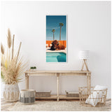 Tall rectangular framed canvas showing a bison relaxing by a turquoise pool with a bright orange wall and two palm trees against a clear blue sky.